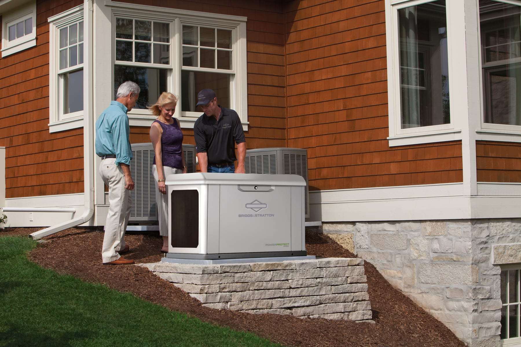 A technician showing two homeowners a Briggs & Stratton PowerProtect standby generator installed on a pad beside their house
